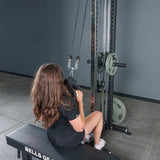 A person with long brown hair, in a black shirt and gray shorts, sits on a workout bench using the Bells of Steel Fat Bar - T-Bar Row Cable Attachment for a lat pulldown, with weight plates loaded on the machine to build grip strength.