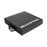 The Bells of Steel Trifold Mat is a black, square, non-slip exercise mat with handles and "BELLS OF STEEL" printed in white on the top.