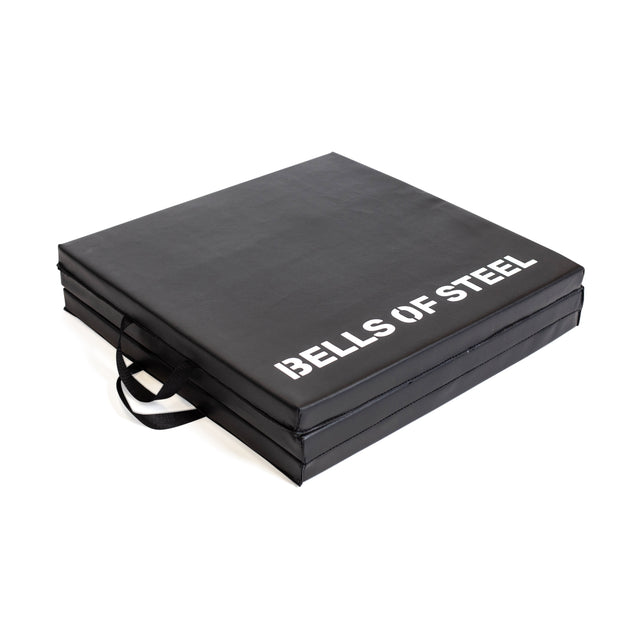 The Bells of Steel Trifold Mat is a black, square, non-slip exercise mat with handles and "BELLS OF STEEL" printed in white on the top.