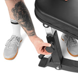 A person with a lion tattoo on their forearm adjusts the knob on a Bells of Steel Adjustable Utility Bench, wearing white socks and gray sneakers with gum soles.