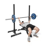 A person in a gray shirt and black shorts performs a bench press with blue weight plates on the Bells of Steel Adjustable Utility Bench, which has a 575lb max capacity.