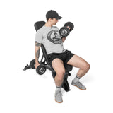 A man in a cap, gray t-shirt, black shorts, and gray shoes sits on the Bells of Steel Adjustable Utility Bench (575lb max capacity), performing bicep curls with dumbbells against a white background.