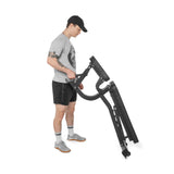 A man in athletic wear and a cap stands on a white background, lifting one end of the Bells of Steel Adjustable Utility Bench to display its versatile incline angles and included bench attachments.