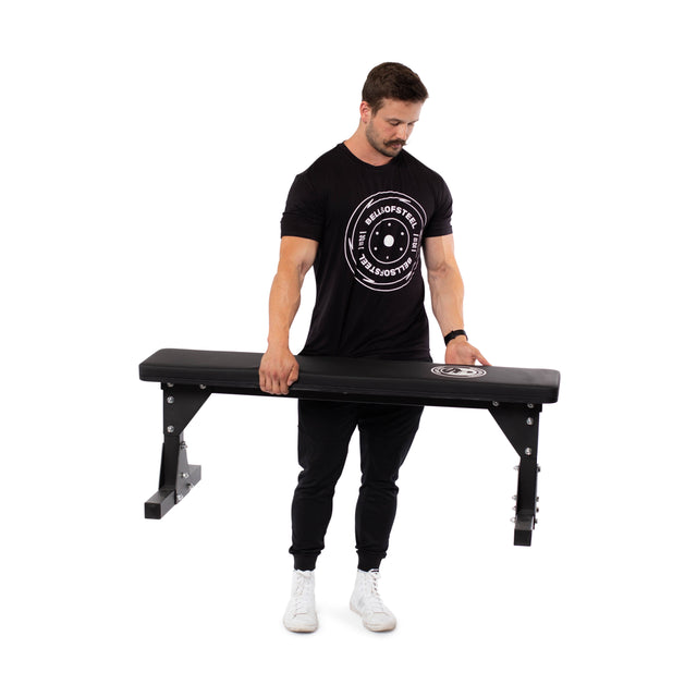 A man in a black t-shirt and pants stands on a white background, effortlessly lifting the Bells of Steel Flat Utility Bench with one hand.