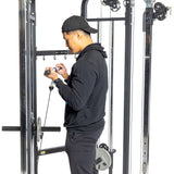 A man in black athletic wear and a cap performs cable curls using the Bells of Steel Functional Trainer. The machine, featuring weight stacks, allows him to focus on his exercise against the plain white background.