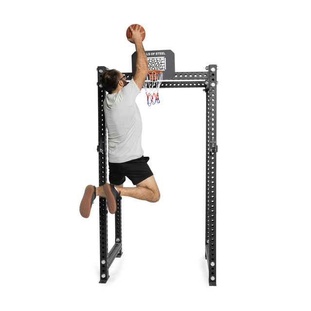 A man in a gray shirt and black shorts jumps to dunk a basketball into the Bells of Steel Rack Attached Basketball Hoop w/ Ball, mounted on a metal frame.