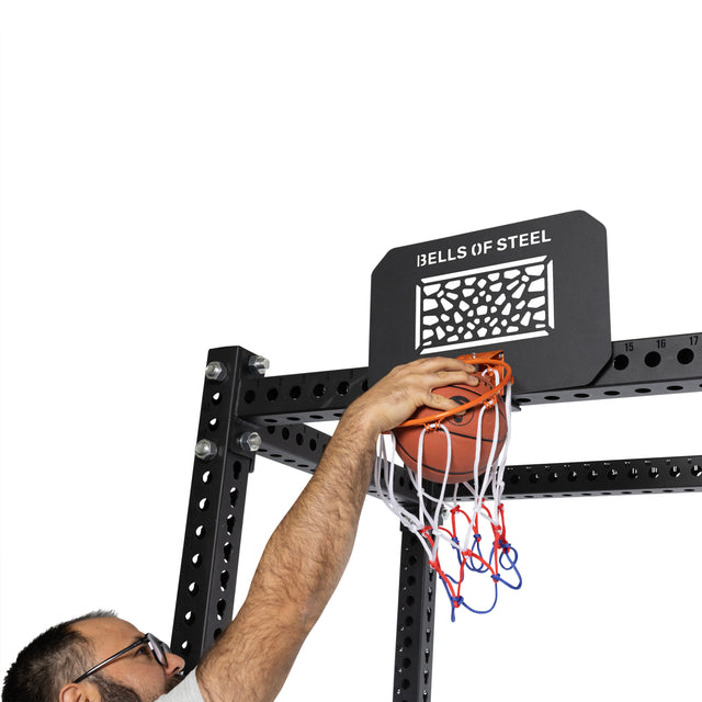 A person wearing glasses dunks a basketball into the Bells of Steel Rack Attached Basketball Hoop w/ Ball, bringing basketball fun to their home gym workout space.