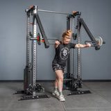 A woman performs a chest press in the gym using the Bells of Steel Lever Arms Rack Attachment, with weights attached. She is dressed in a black t-shirt, printed shorts, socks, and athletic shoes.