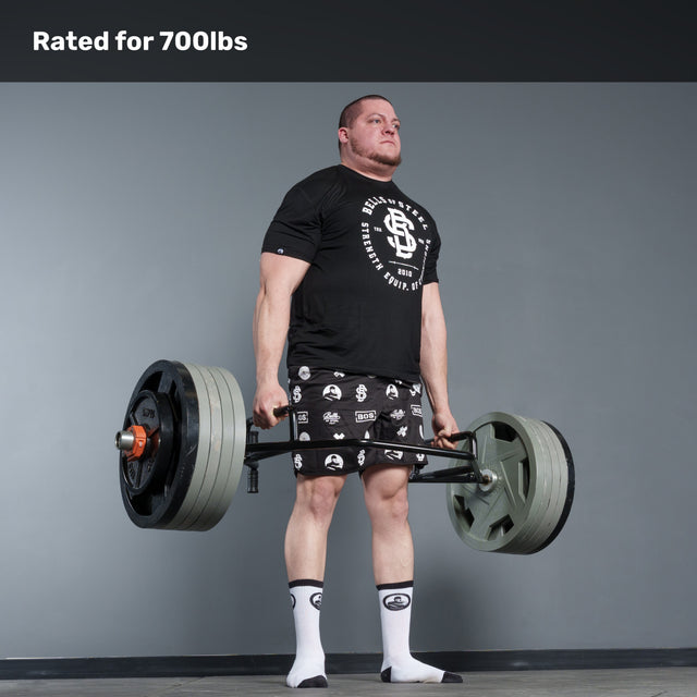 A man in a black t-shirt, patterned shorts, and socks lifts a heavily loaded Bells of Steel Open Trap Bar / Hex Bar in a gym. Text above reads "Rated for 700lbs." The background is plain gray.