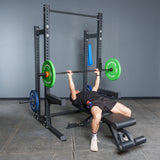 A person in a gym is lying on a bench, performing a bench press with a barbell loaded with green and orange plates inside the Bells of Steel Hydra Half Rack - Prebuilt (3" x 3", ⅝" Holes).