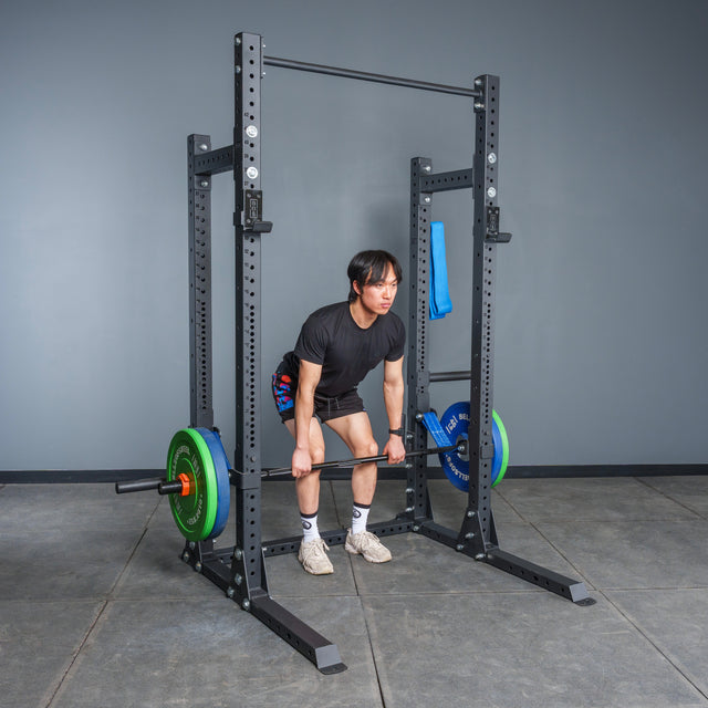 A person in a black t-shirt and shorts is deadlifting with a loaded barbell inside the Bells of Steel Hydra Half Rack - Prebuilt (3" x 3", ⅝" Holes) in a gym with concrete floors and a plain gray wall.