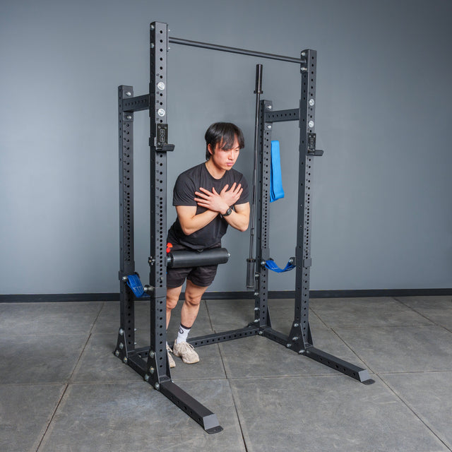 A person does a Nordic hamstring curl in the Bells of Steel Hydra Half Rack - Prebuilt (3" x 3", ⅝" Holes), ankles secured by a padded bar and arms crossed, leaning forward in a gym.