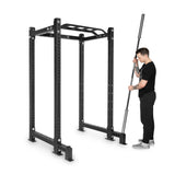 A man in black clothes holds a barbell next to a Bells of Steel power rack with Front Foot Stabilizers and a pull-up bar attachment on top.