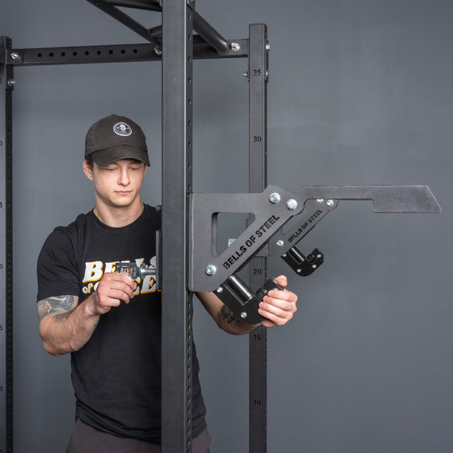 A man in a black Bells of Steel shirt and cap adjusts the Monolift Rack Attachment by Bells of Steel on a squat rack against a gray background.