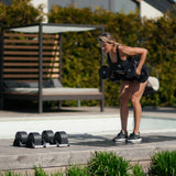 A woman in workout clothes uses Nuobell NÜOBELL Adjustable Dumbbells to perform a bent-over row on a wooden deck outdoors, with extra weights and a lounge chair visible in the background.