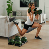 Wearing workout clothes, a woman does a Bulgarian split squat in her living room with Nuobell NÜOBELL Adjustable Dumbbells, resting her back foot on the couch while more dumbbells lie on the floor nearby.