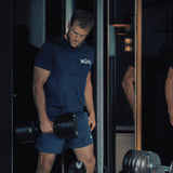 A man in a navy blue athletic outfit stands by a mirror at the gym, holding a Bells of Steel NÜOBELL-S Adjustable Dumbbell, looking down with focus. Various gym equipment is visible in the background.
