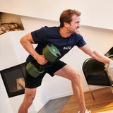 A man in a navy T-shirt and black shorts works out indoors, lifting a Nuobell NÜOBELL Adjustable Dumbbell with one arm while bracing against a chair, looking focused and determined.