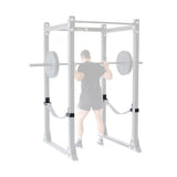 Wearing athletic gear, a person performs a barbell squat inside a rack equipped with Bells of Steel Safety Strap Connectors. The rack’s uprights are subtly faded to draw focus to the lifter.