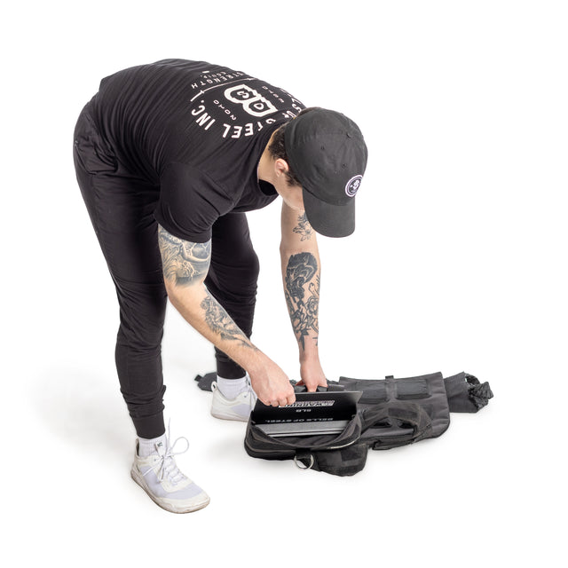 Dressed in black athletic gear and a cap, an individual with visible arm tattoos places a Bells of Steel Weighted Vest and Plates into a black backpack against a white background.
