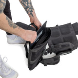 A person with tattooed arms adds a metal plate to the Bells of Steel Weighted Vest and Plates, shown open on a white background. This workout accessory offers an adjustable fit. Only their lower body and hands are in view.